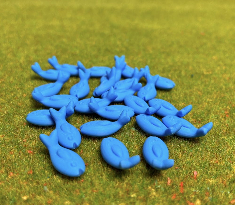 A school of small blue 3D-printed fish tokens scattered across a green board game mat